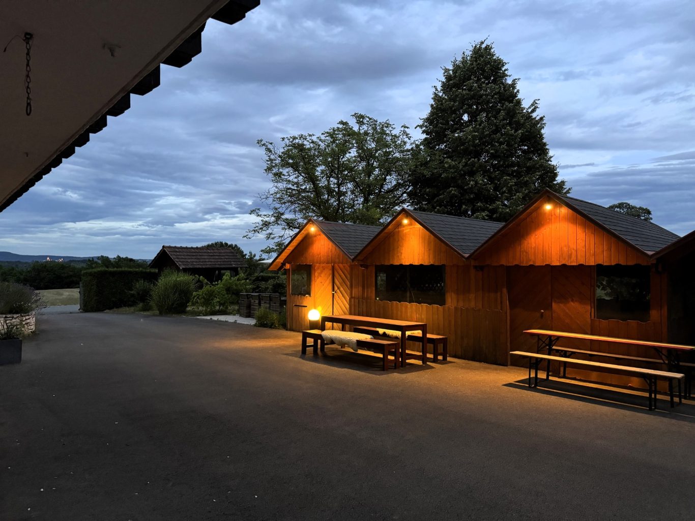 Holzhütten mit Beleuchtung bei Dämmerung, umgeben von Bäumen und Wolken am Himmel.